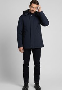 Chaqueta azul marino con capucha, cremallera frontal, dos bolsillos laterales y textura suave. Combinada con vaqueros oscuros y zapatos negros.