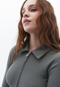 Grauer, gerippter Zip-Up-Oberteil mit Kragen und silbernem Reißverschluss, präsentiert mit schulterlangen, welligen braunen Haaren vor einem weißen Hintergrund.