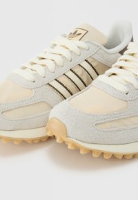 Baskets Adidas beige et gris clair avec détails en daim, lacets blancs, les trois bandes emblématiques et semelles en gomme texturées.