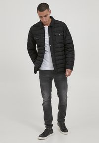 Sort puffet jakke med quiltet tekstur, trykknapper og to brystlommer, parret med en hvid t-shirt, grå jeans og sorte sneakers.