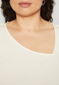 Witte geribde top met een brede, asymmetrische halslijn en subtiele textuur. Beschikt over een gladde stof zonder duidelijke patronen of versieringen.