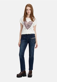 Young woman wearing white T-shirt with leopard print heart, dark blue jeans with embellished pocket, and brown platform boots, standing.