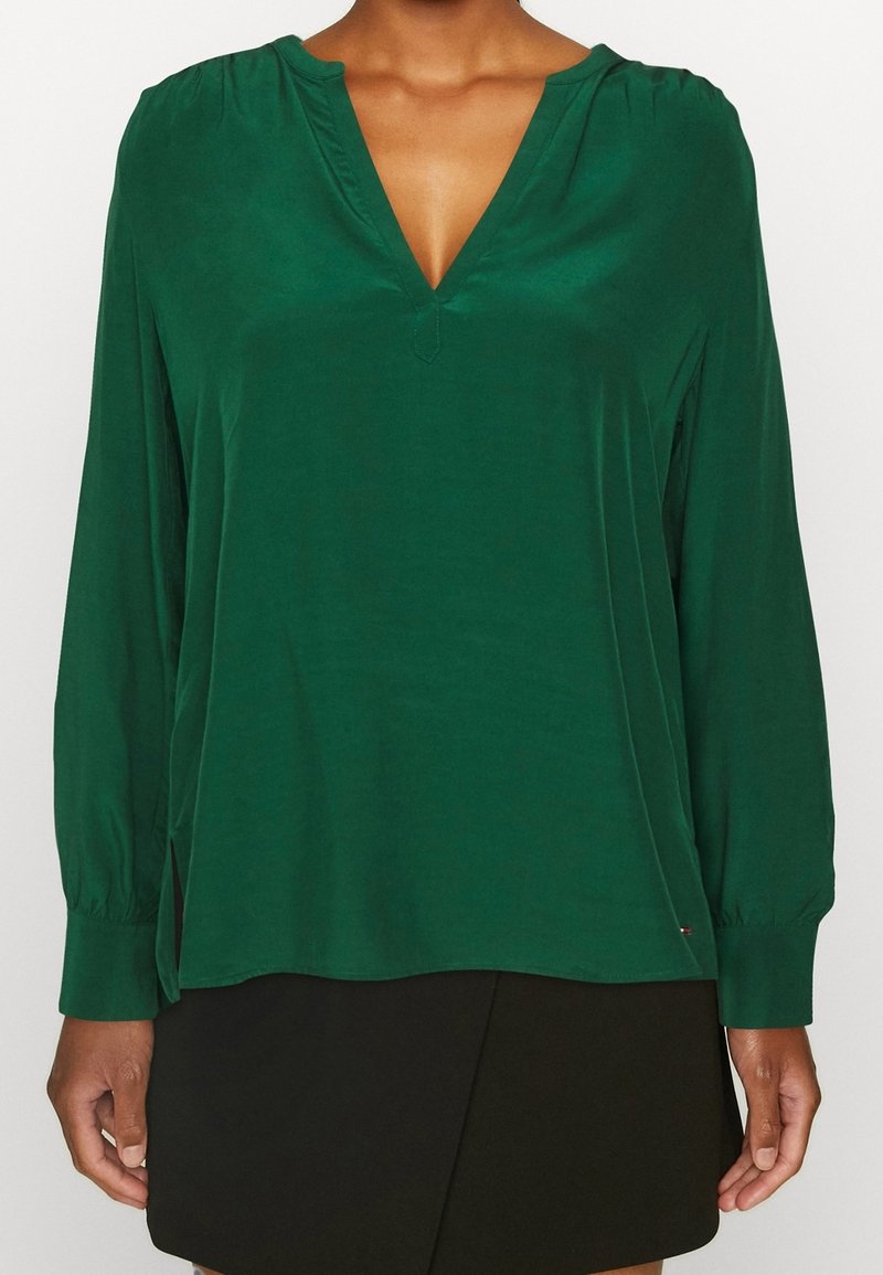 Zielona bluzka z długim rękawem, dekoltem w kształcie litery V i marszczonymi ramionami. Miękka tkanina, boczne szwy oraz detal z logo blisko dołu.
