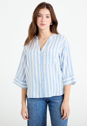 Młoda kobieta w jasnoniebiesko-białej koszuli w pionowe paski z trzeciowązowymi rękawami i niebieskich dżinsach, stojąca na białym tle.