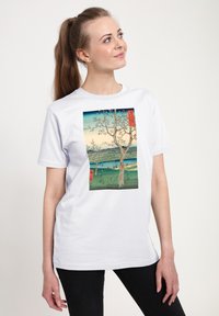 Bílé tričko s potiskem krajiny s stromy, vodou a horami. Design kombinuje odstíny modré, zelené a růžové.