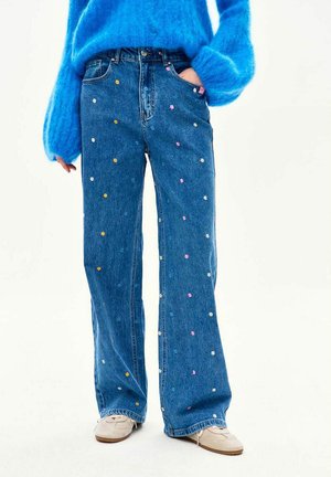 Osoba ubrana w niebieskie, szerokie spodnie dżinsowe z wysokim stanem, ozdobione małymi, wielokolorowymi haftowanymi kwiatami, niebieski puszysty sweter oraz beżowe sznurowane buty.