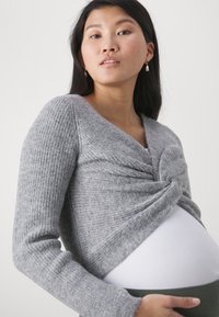 Lichtgrijze gebreide cardigan met een geknoopte voorkant en lange mouwen, gecombineerd met een witte top en groene onderkant. Zachte textuur is duidelijk zichtbaar.