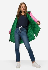 Vaatetakki, jossa on vihreä ja vaaleanpunainen päällinen, kontrastoiva sisus, vetoketjukiinnitys ja huppu, yhdistettynä sinisiin farkkuihin ja valkoisiin lenkkareihin.