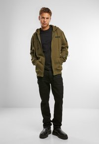 Felpa verde oliva con zip e tasche sopra una maglietta nera; abbinata a jeans neri e stivaletti. Colori solidi e design semplice.