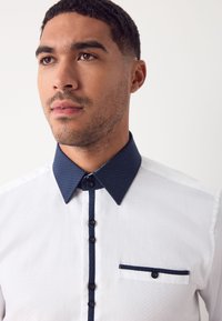 Camicia bianca con tessuto strutturato, caratterizzata da colletto e patta dei bottoni blu scuro, arricchita da una piccola tasca sul petto e dettagli a contrasto.