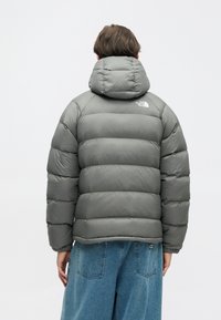 Szürke puffmellény kapucnival, tűzött részekkel és egy kis logóval a hátán. Az anyag sima, fényes textúrájú.