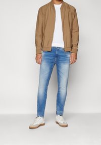 Benvit bomberjacka, vit t-shirt, ljusblå slim-fit jeans och vita sneakers med beige detaljer, stående mot en neutral bakgrund.