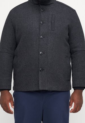 Veste en laine gris foncé boutonnée avec poches latérales portée par une personne à la peau foncée et un pantalon bleu marine, visible du cou aux hanches.