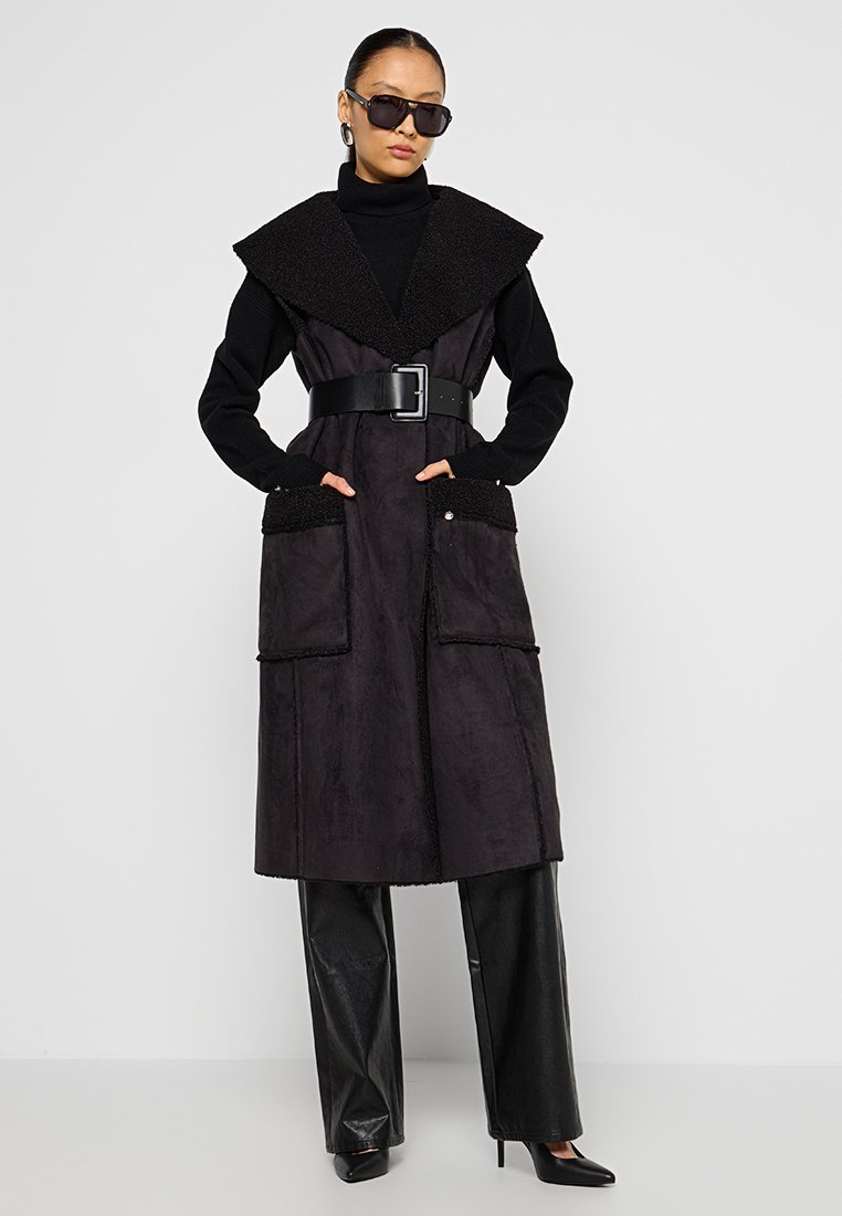 Schwarzer Trenchcoat mit strukturiertem Kragen, tailliertem Bund und großen Taschen. Trägt dazu hochgeschnittene Hosen mit glänzender Oberfläche und Sonnenbrille.