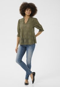 Olive krāsas mežģīņu blūze ar V-veida kakla izgriezumu un trim ceturtdalītām piedurknēm, kombinēta ar šaurām zilām džinsām un melniem balerīnu apaviem, rotāta ar ziedu rakstu.