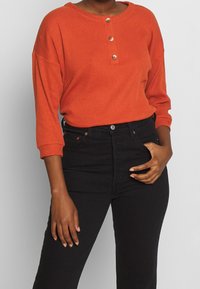 Ribbat, orange långärmad topp med tre knappar vid halsringningen, parat med högmidjade svarta jeans. Framhäver textur och färgkontrast.