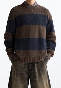 Pull en tricot avec rayures marron et bleu marine, présentant une texture côtelée, un col rond et des épaules tombantes, associé à un pantalon foncé délavé.