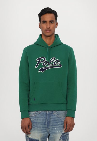 Grønn hettegenser med lomme foran, stort "Polo"-logo i hvitt og marineblått, og ribbestrikkede mansjetter. Bæres med slitte blå jeans.