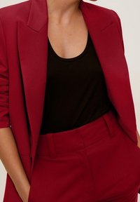 Persoon in een dieprode blazer en bijpassende high-waisted broek met een zwarte top met scoopneck, handen in de zakken, tegen een neutrale achtergrond.