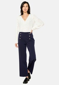 Blusa bianca in pizzo con bottoni frontali e maniche a sbuffo, abbinata a pantaloni blu navy a vita alta con dettagli in bottoni dorati e gambe larghe.