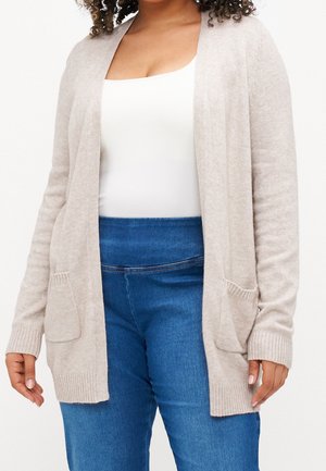 Femme portant un cardigan beige ouvert avec des poches par-dessus un haut blanc et un jean bleu taille haute, visible des épaules aux hanches.