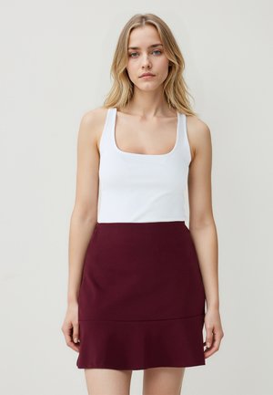 Une femme blonde portant un haut sans manches blanc et une jupe courte bordeaux se tient devant un fond clair uni.