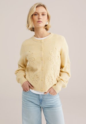 Femme blonde portant un cardigan jaune boutonné avec des motifs floraux en relief, chemise blanche en dessous, jeans bleu clair, mains dans les poches.