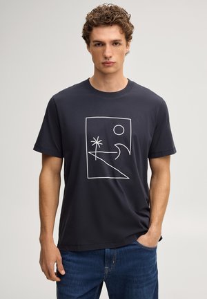 Junger Mann mit lockigem Haar trägt ein schwarzes T-Shirt mit weißem abstraktem Landschaftsdesign und blaue Jeans, steht vor einem schlichten Hintergrund.