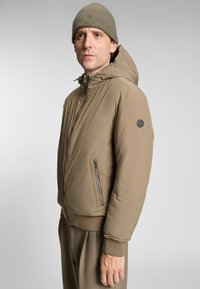 Giubbotto imbottito marrone chiaro con cappuccio, dotato di una tasca con cerniera e polsini a costine. Abbinato a pantaloni marroni coordinati e un berretto a maglia.