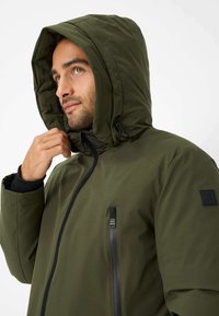 Giacca verde con cappuccio, dal tessuto liscio, chiusura con zip e tasca laterale. Presenta una toppa con logo sulla manica.