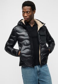 Άνδρας που φοράει μαύρο γυαλιστερό μπουφάν puffer με μπεζ επένδυση, μαύρο πουκάμισο και μπλε τζιν, τα χέρια στις τσέπες, κοιτάζοντας προς το πλάι.