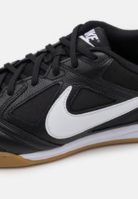 Juodas sportinis batelis su tekstūruota oda ir tinklo viršutine dalimi. Yra baltas Nike ženklas, juodos raištelės ir guminis padas su guma akcentu.