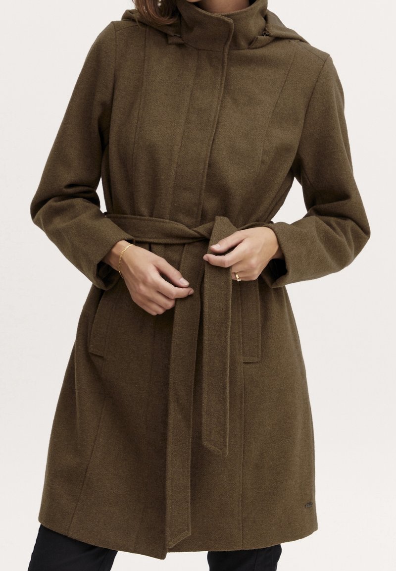 Kvinde, der spænder et olivengrønt uldfrakkekned længere ned til knæet med et bælte, stående mod en ensfarvet lys baggrund.