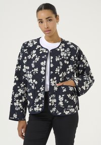 Quilted navy jacket with white floral pattern, round neck, no closure, and two front pockets. Smooth fabric with contrasting trim.
