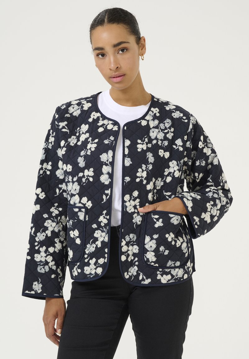 Quilted navy jacket with white floral pattern, round neck, no closure, and two front pockets. Smooth fabric with contrasting trim.