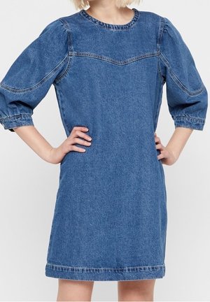 Frau trägt ein knielanges blaues Jeanskleid mit dreiviertellangen Puffärmeln und hat die Hände in die Hüften gestützt vor einem einfarbigen Hintergrund.