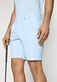 Svetlemodré kraťasy z texturované tkaniny, s bočními sklady, elegantním designem a čistým lemem. Model drží v ruce golfovou hůl.