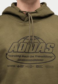 Gros plan d'un sweat à capuche Adidas vert olive avec un lacet noué en nœud papillon et un texte noir promouvant des services de skateboard.