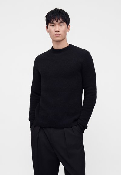 Junger Mann, der einen schwarzen Langarmpullover und schwarze Hose trägt, steht mit den Händen in den Taschen vor einem einfarbig hellen Hintergrund.