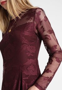 Burgundské krajkové šaty s dlouhými rukávy, květinovými vzory, texturovanou tkaninou a přiléhavým korzetem s rozšířenou sukní.