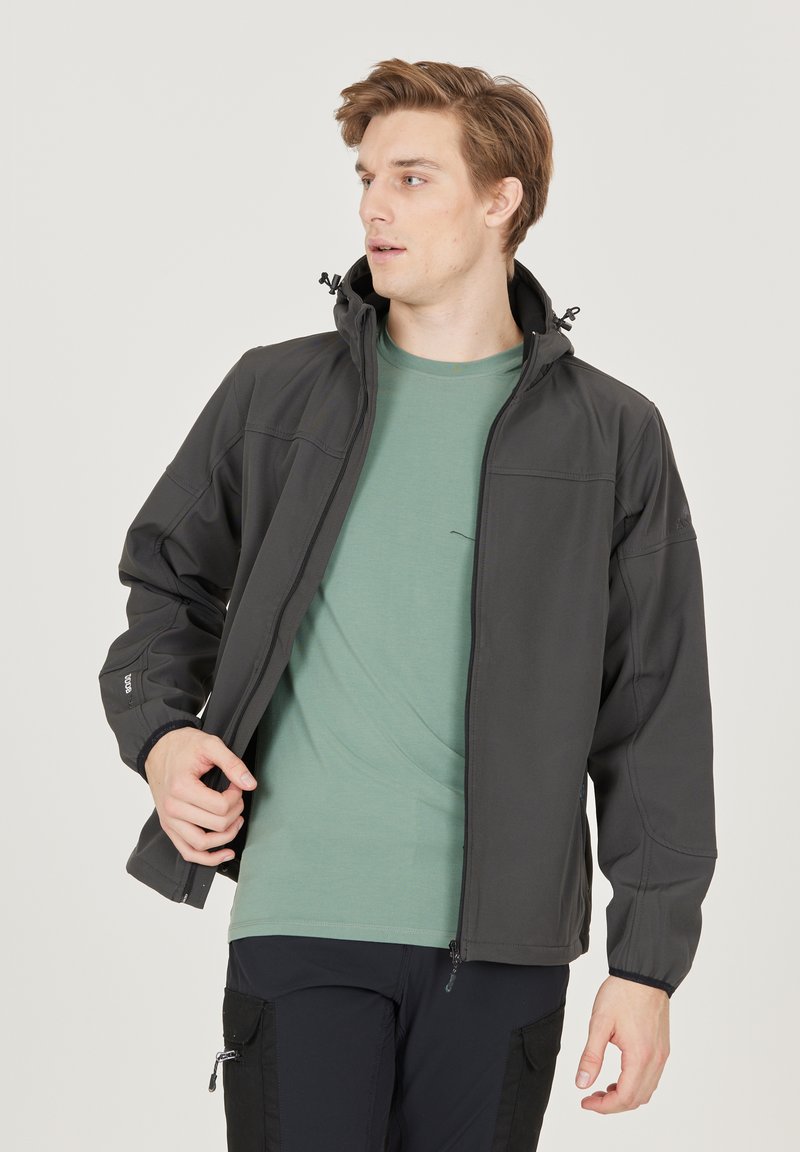 Herremodel sort softshell-jakke med lynlås fortil, justerbar hætte, elastiske manchetter og glat tekstur. Båret over en grøn t-shirt.