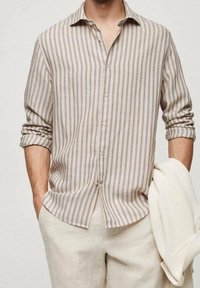 Chemise à rayures beige et marron clair avec boutons, à coupe décontractée, manches longues et texture douce. Associée à un pantalon clair.