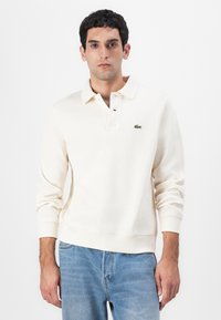 Krämfärgad långärmad polo skjorta i ribbad textur, med en treknapplåsning och en grön broderad logotyp på vänster bröst.