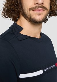 Marinefarbener Baumwoll-T-Shirt mit kurzen Ärmeln, Rundhalsausschnitt und einem dekorativen Schulterüberwurf. Enthält ein horizontal angebrachtes Logoband.