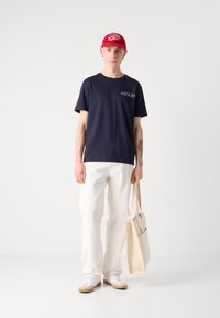 Marineblaue Baumwoll-T-Shirt mit Logo, weiße lässige Hose, beigefarbene Tragetasche, weiße Sneakers mit beigen Akzenten und rote Baseballkappe.