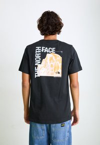 Sort bomulds T-shirt med et stort grafik af et bjerg på ryggen. Accentueret med hvid og orange tekst. Løs pasform, rund halsudskæring.