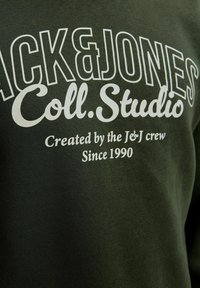 Mørkegrøn stof med hvid tekst, der læser "JACK & JONES Coll. Studio Skabt af J&J-crew siden 1990" i forskellige fonte.