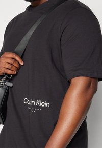 Mies, joka pukeutuu mustaan Calvin Klein -t-paitaan, jossa on valkoinen logo, ja kantaa mustaa nahkaluukkua leveällä hihnalla olkapäillään.