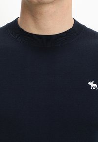 Granatowy sweter dziany z okrągłym dekoltem. Na lewej piersi subtelne, białe logo łosia. Gładka tekstura, bez widocznych wzorów.