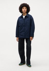 Giacca blu navy, con bottoni e colletto, dotata di tasca frontale, abbinata a jeans scuri e scarpe verdi, in piedi su uno sfondo chiaro.